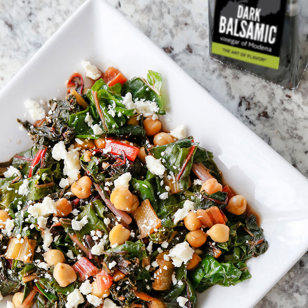 Mediterranean Swiss Chard made with Caramelized Garlic Infused Olive Oil, Sun Dried Tomato Balsamic Vinegar and Roasted Garlic Sea Salt served in a white square dish atop a marble table.