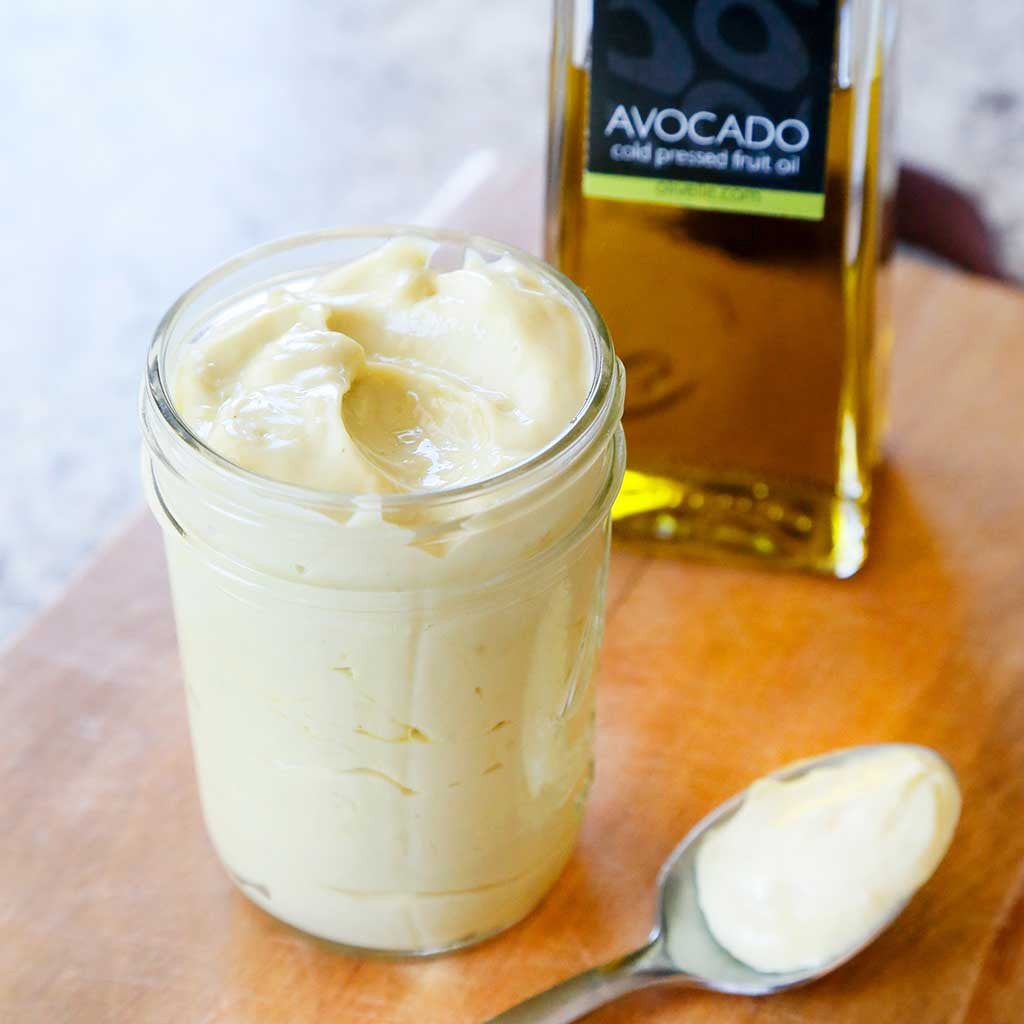 avocado oil mayonnaise in mason jar with spoon alongside avocado oil bottle on a wooden board