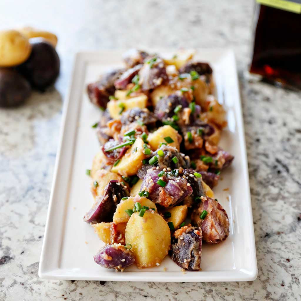 Salt & Vinegar Potato Salad is a combination of Prosecco whole grain mustard, vinegar, oil, bacon and topped with chives served on a white platter next to a bottle of Spanish sherry vinegar