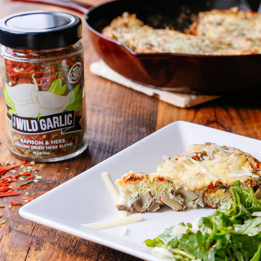 Wild Garlic & Mushroom Frittata (Baked Eggs) with Wild Garlic Dried Herb Blend, Caramelized Garlic Infused Olive Oil, Smokey Bacon Sea Salt, Crimini Mushrooms and Parmesan Cheese served on a white plate next to a green salad.