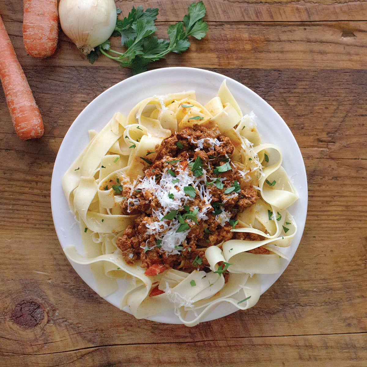 Pappardelle pasta with Bolognese sauce and grated cheese on a white plate on a wooden table, with carrots and an onion beside it