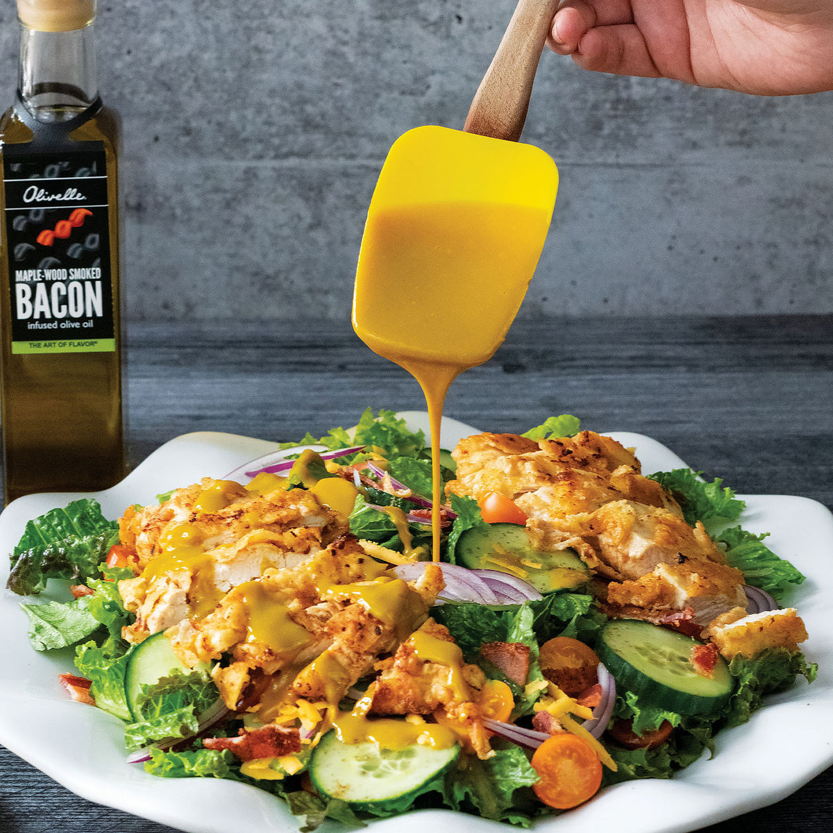 Golden dressing being drizzled over a kale and vegetable salad on a white platter, with a bottle of Maple-Wood Smoked Bacon Infused Olive Oil behind