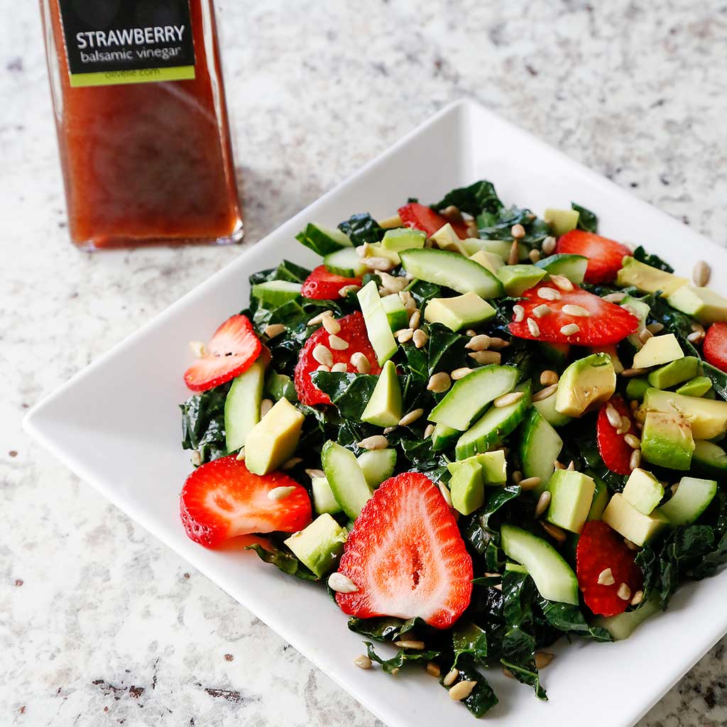 Strawberry kale salad with sliced cucumber and avocado on a white square plate, with a bottle of vinegar behind