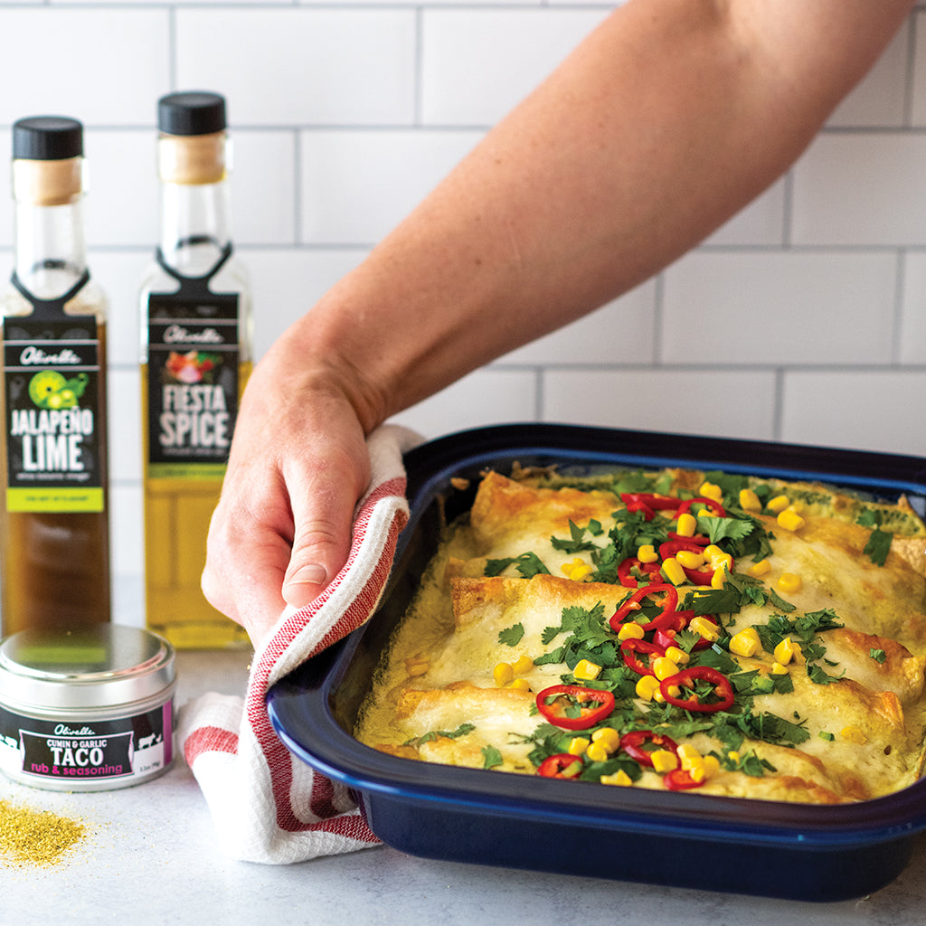 Baked white chicken enchiladas topped with corn and peppers in a navy baking dish held with an oven mitt, with bottles and a jar of seasoning behind