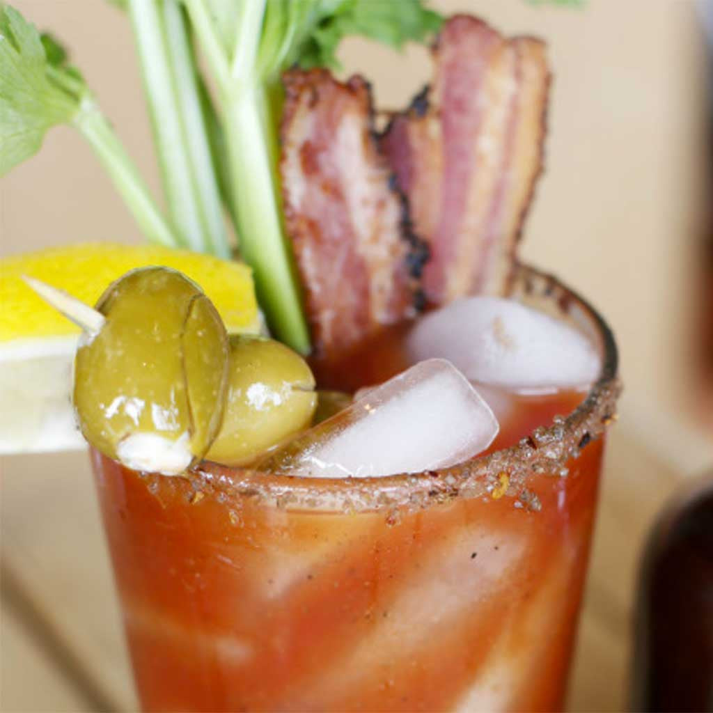 A Bacon Bloody Mary with a slice of lemon, stuffed olives, cooked bacon pieces, and a stalk of celery served in a glass rimmed with smokey bacon sea salt