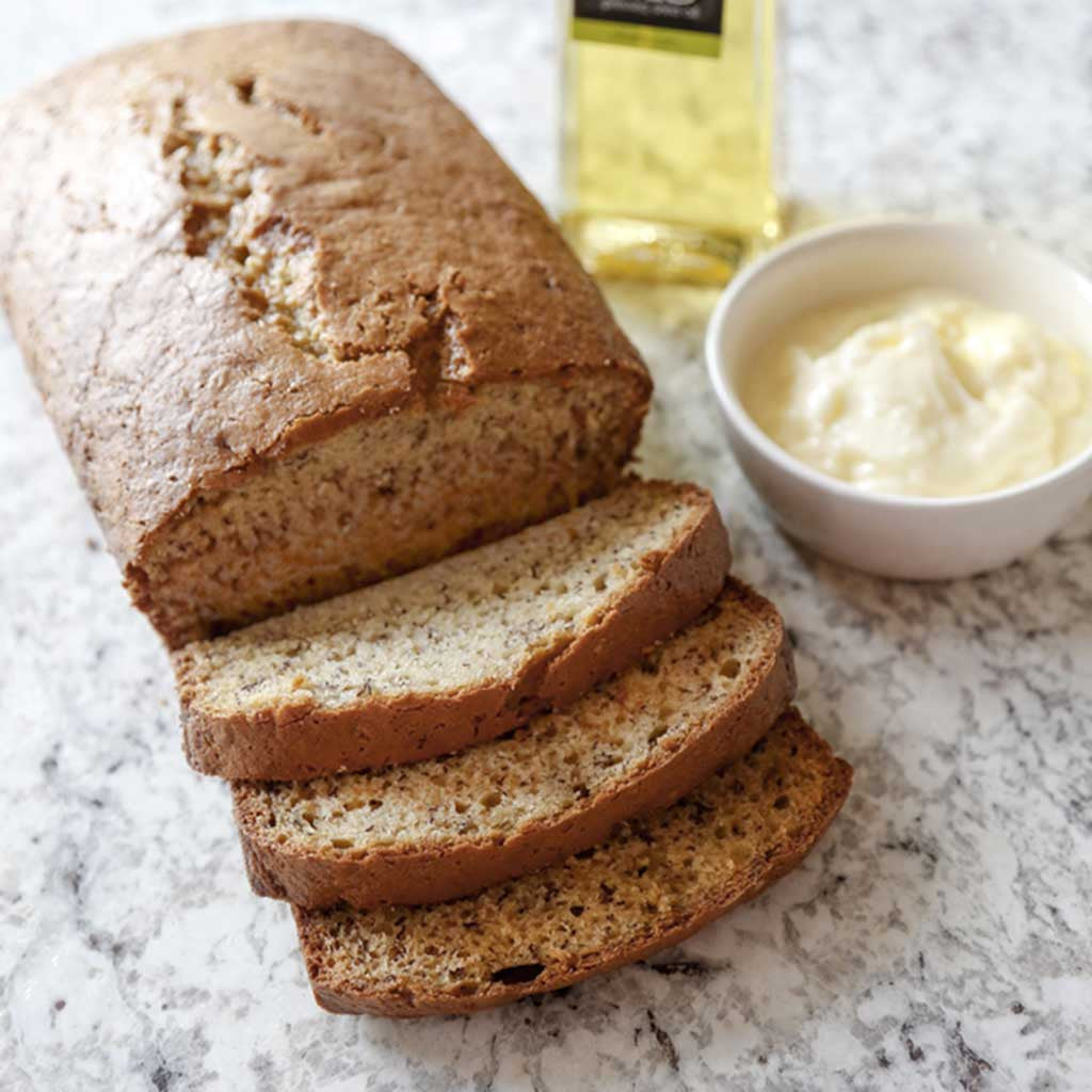 A loaf of banana bread half sliced next to a bowl of whipped olive oil butter and a bottle of Extra Virgin Olive Oil