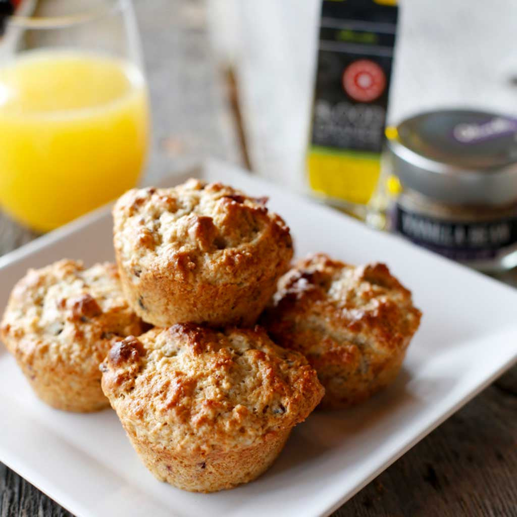 A white plate of Cranberry and Pecan muffins made with Blood Orange Olive Oil and Vanilla Bean Sea Salt served next to a glass of orange juice.