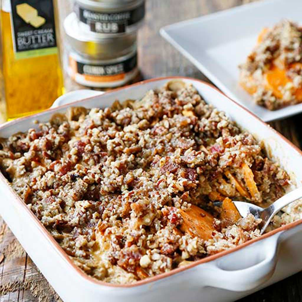 A warm bourbon sweet potato gratin with bacon pecan crumble served in a white baking dish next to a jar of seasoning, sea salt and a bottle of sweet butter olive oil