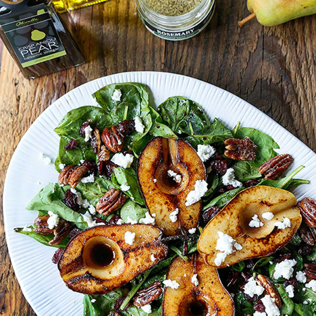 A caramelized pear and pecan salad topped with feta or cheese served on a white platter next o a bottle of pear balsamic vinegar and a tin of seasoning