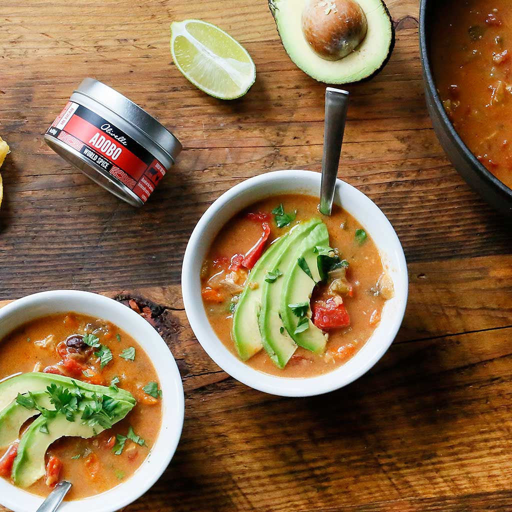 Creamy Mexican Chicken Soup topped with fresh cilantro, avocado slices, tomatoes, and a slice of lime served in white bowls on a rustic wood board next to a tin of Abodo seasoning