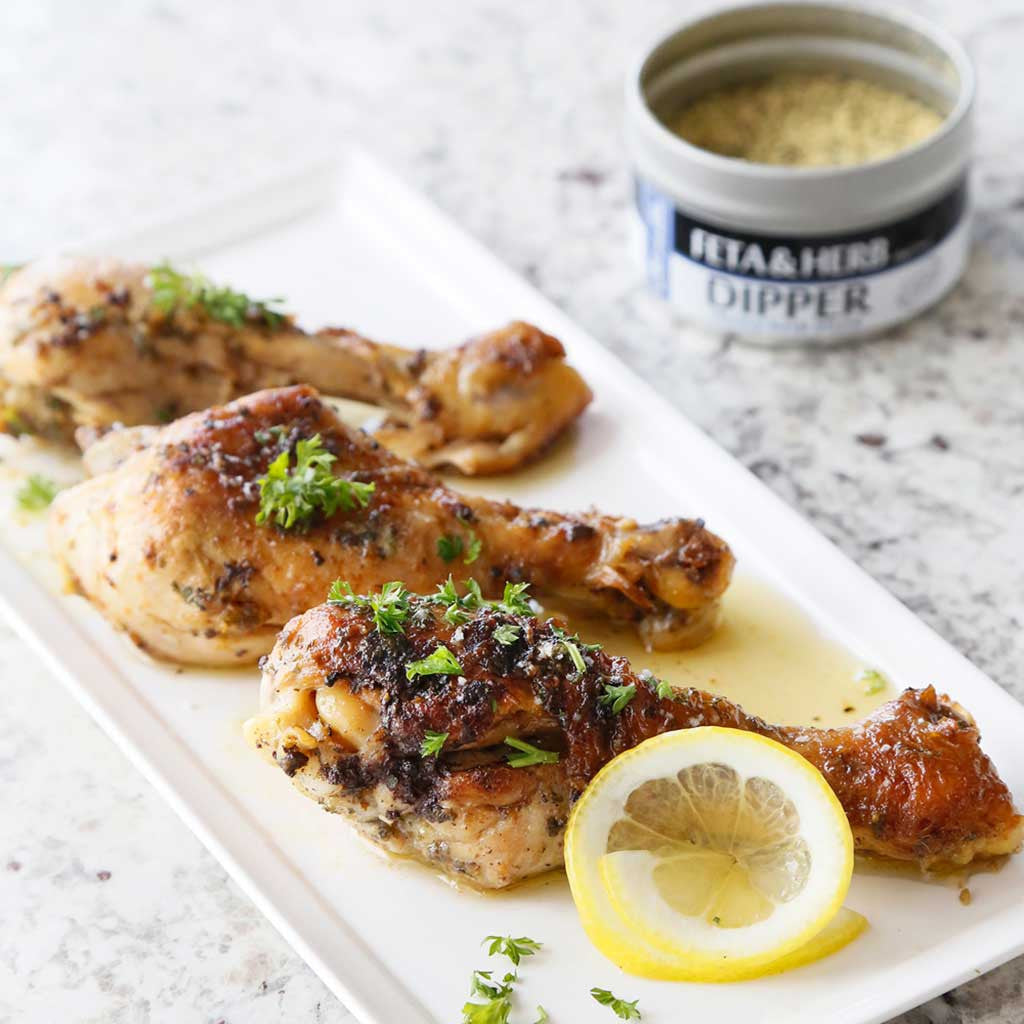 Chicken drumsticks seasoned with Feta and Herb Dipper and Citrus Dill Sea Salt, cooked with Sicilian Lemon or Caramelized Garlic Olive Oil, and sprinkled with parsley served  on a white plate next to a tin of feta & herb dipper