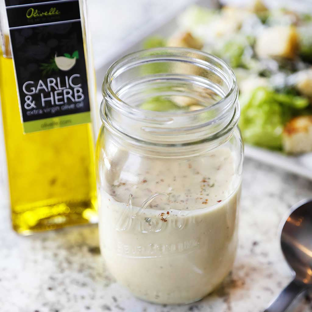 A creamy, garlicky, and fresh tasting salad dressing using Avocado Oil, Garlic and Herb Oil, vinegar, sea salt, and herb seasoning served in a mason jar next to a Caesar salad and bottle of  olive oil
