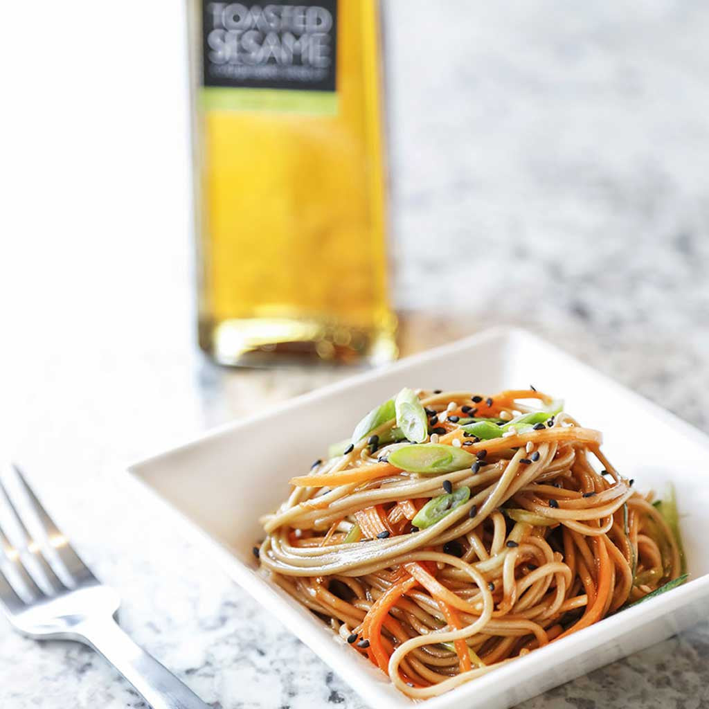 garlic sesame soba noodle salad in white square dish with a fork alongside toasted sesame oil served on a marble table top