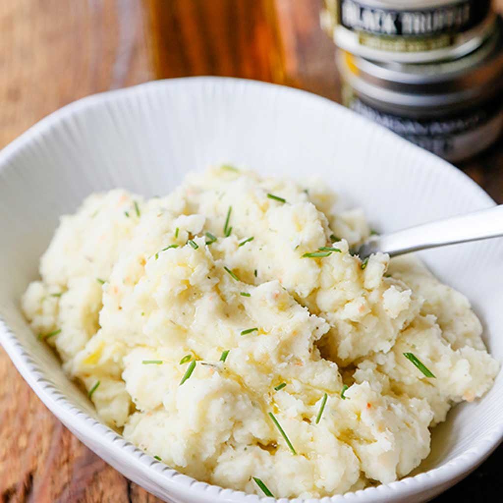A bowl of mashed potatoes topped with Parmesan cheese and truffle olive oil served next to a tin of black truffle sea salt