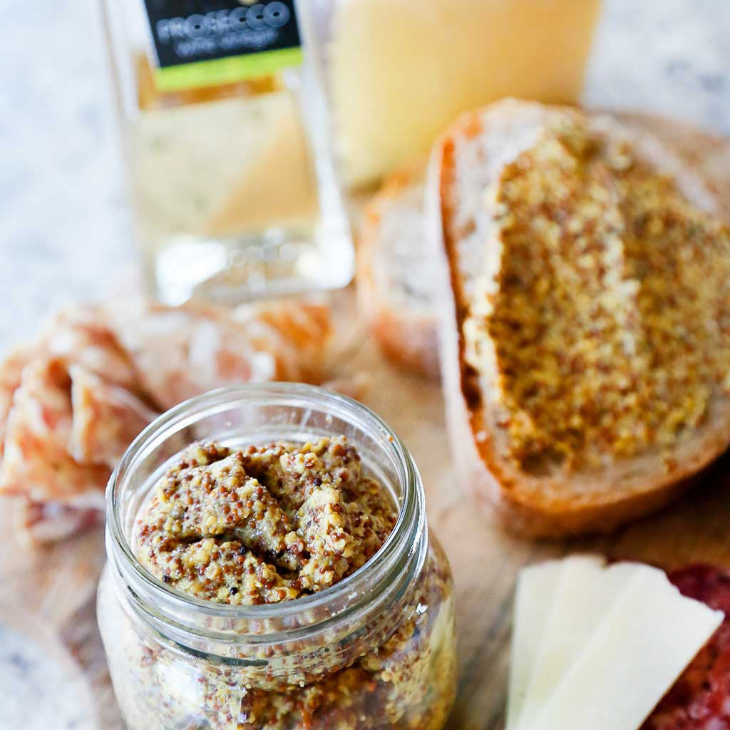 Prosecco Whole Grain Mustard made with Prosecco sparkling wine,  Prosecco wine vinegar, brown/yellow mustard seeds and sea salt served in a mason jar next to a slice of bread topped with the mustard spread and a bottle of prosecco vinegar