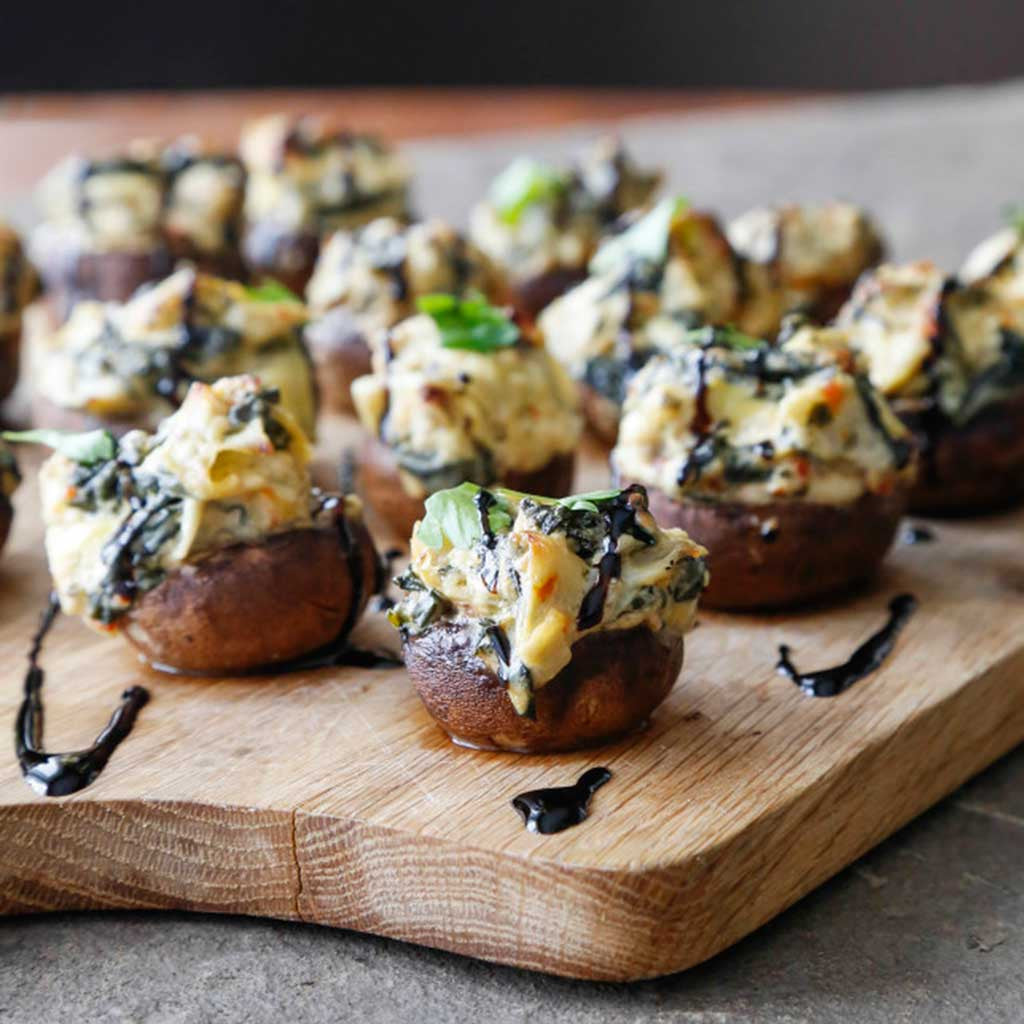 Spinach and Artichoke Stuffed Mushroom Bites drizzled with balsamic glaze served on a wooden cutting board.
