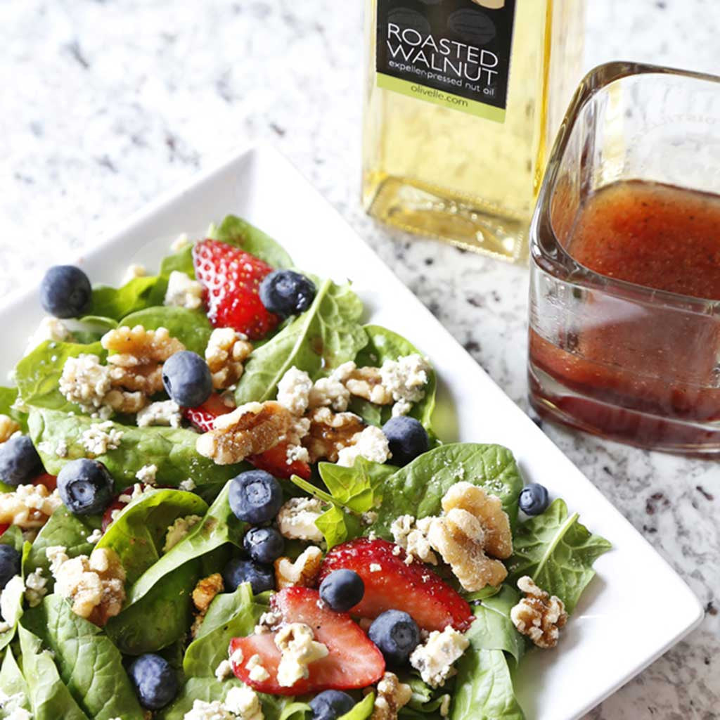 Spinach walnut berry salad on a white plate served next to a jar of roasted walnut oil and raspberry balsamic vinegar