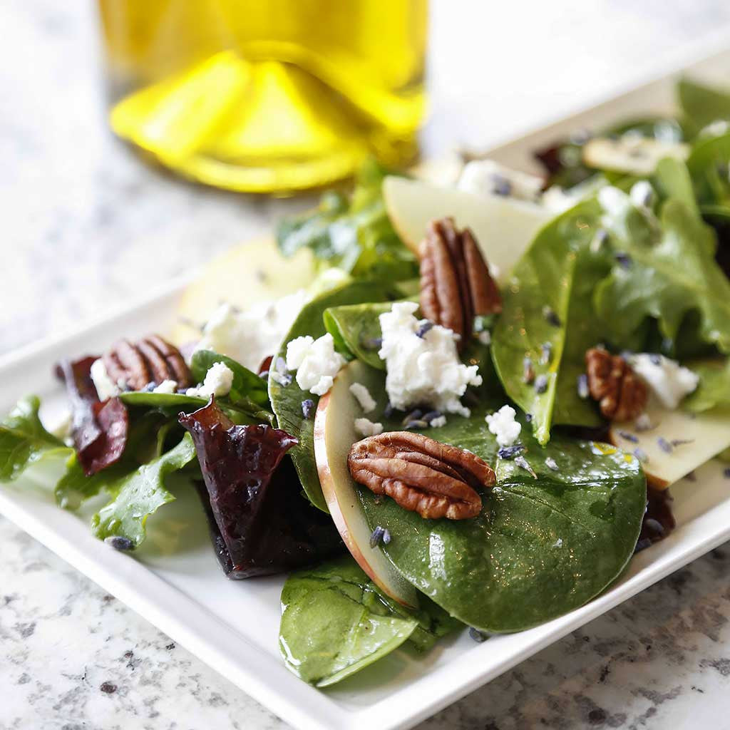 A refreshing salad tossed with greens, apples, pecans, goat cheese, and a lavender vinaigrette dressing served on a white rectangular plate next to a bottle of olive oil.