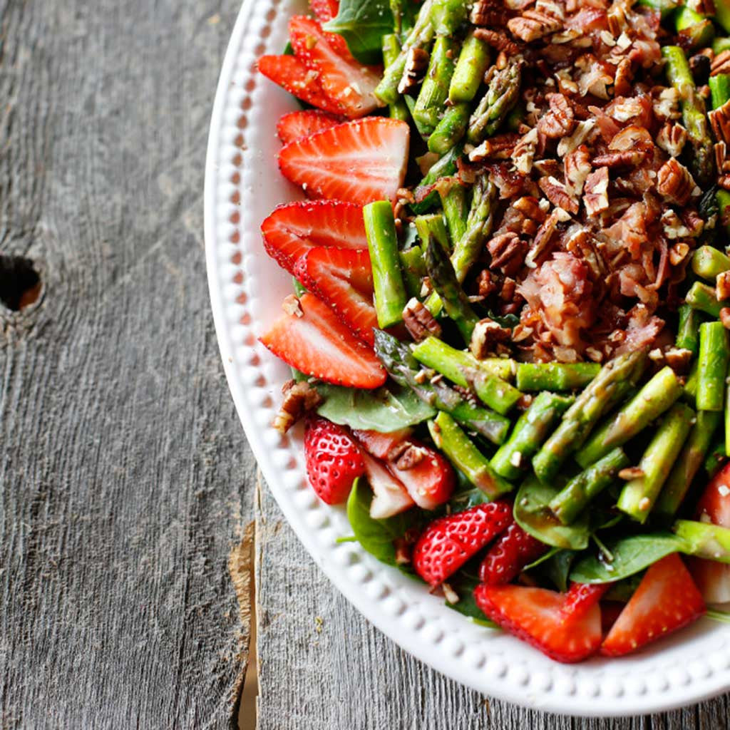 A colorful salad with strawberries, asparagus, drizzled with vinaigrette served in a white platter on a rustic, wood table. 