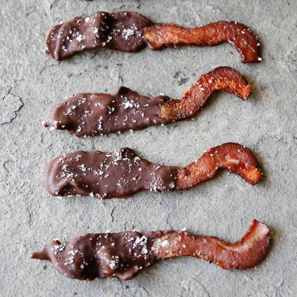 Four chocolate dipped pieces of bacon with sprinkled sea salt atop a concrete counter top