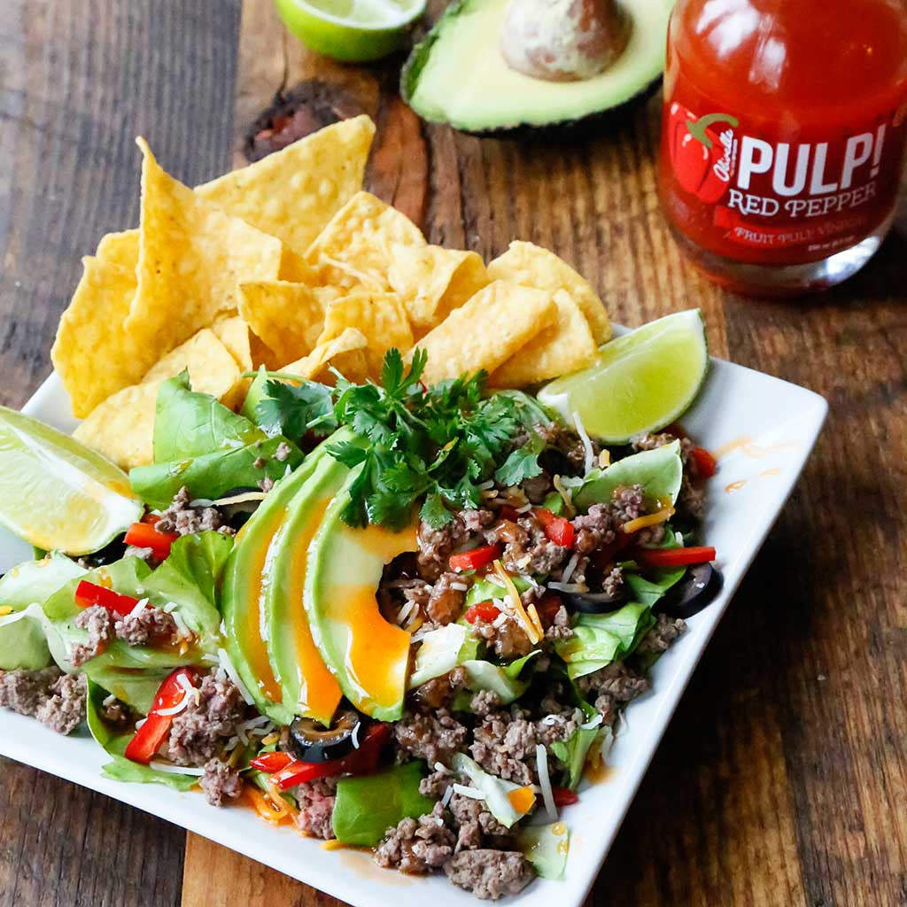 Red Pepper Taco Salad with side of avocados and tortilla chips served on a white square plate on a rustic table top next to avocado halves and Pulp! Red Pepper Fruit Pulp Vinegar
