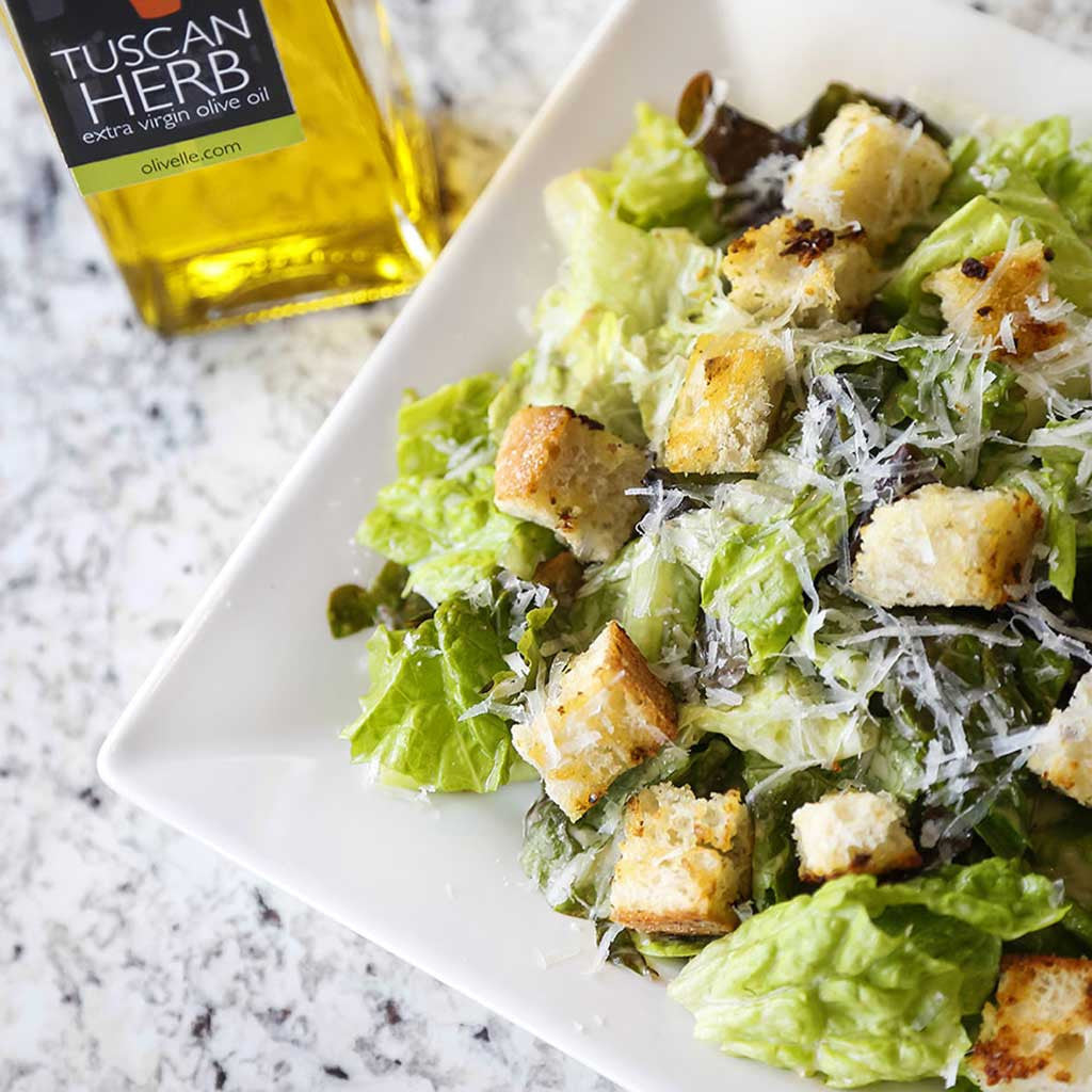 Tuscan Herb Croutons drizzled with Tuscan Herb Olive Oil and sprinkled with our garlic and herb seasoning atop a green Caesar salad served next to a bottle of Tuscan herb olive oil