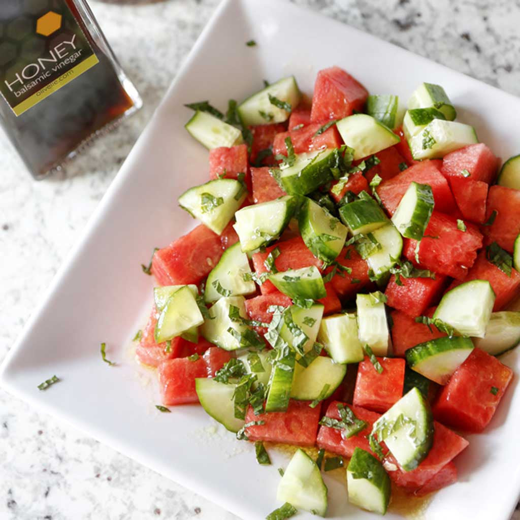 A light and refreshing side dish combining watermelon, cucumbers, mint leaves, our Honey Balsamic Vinegar and Sicilian Lemon or Basil Genovese Olive Oil served on a white salad plate next to a bottle of honey vinegar
