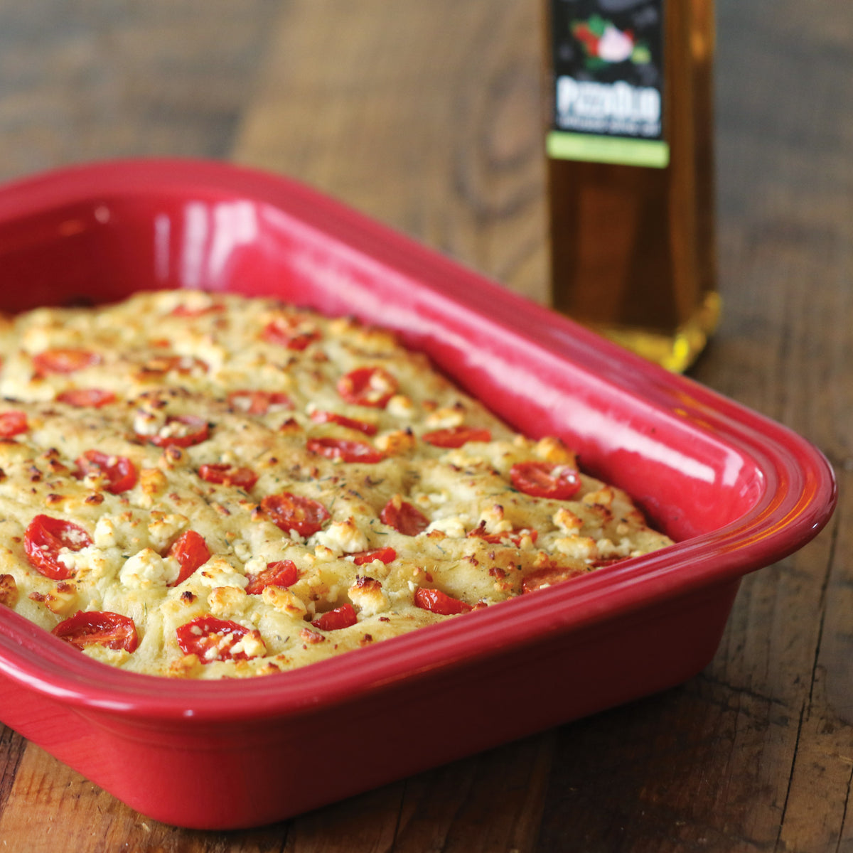 Gluten free focaccia baked in a red dish, topped with cherry tomatoes, with a bottle of olive oil behind