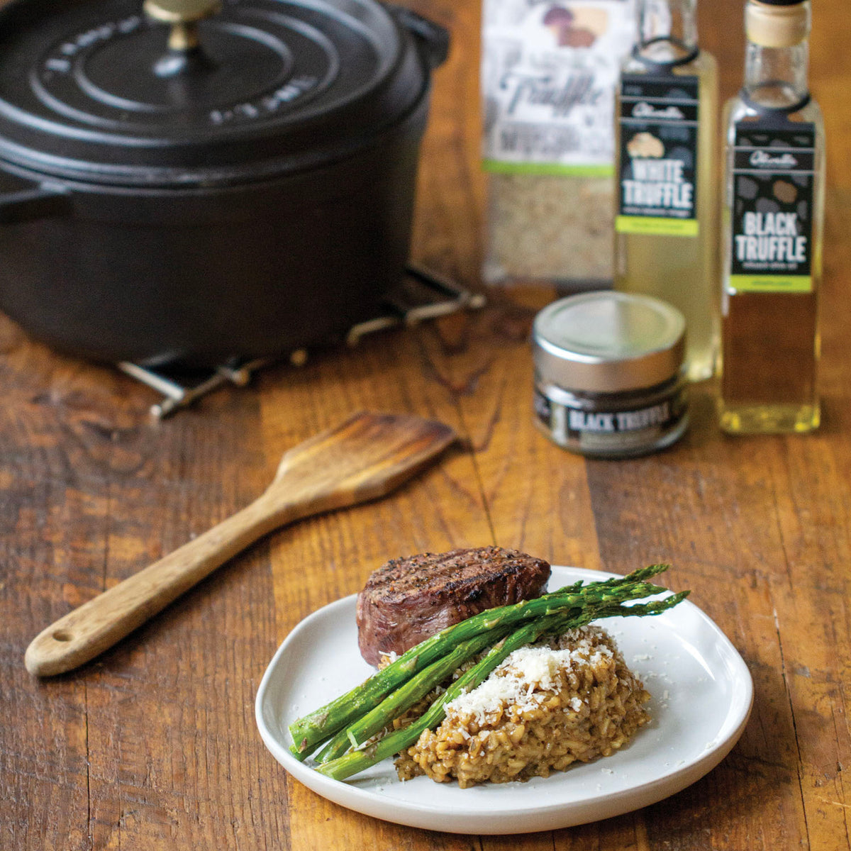 Parmesan truffle wild mushroom risotto beside seared steak and asparagus on a white plate, with a Dutch oven, wooden spoon and bottles behind