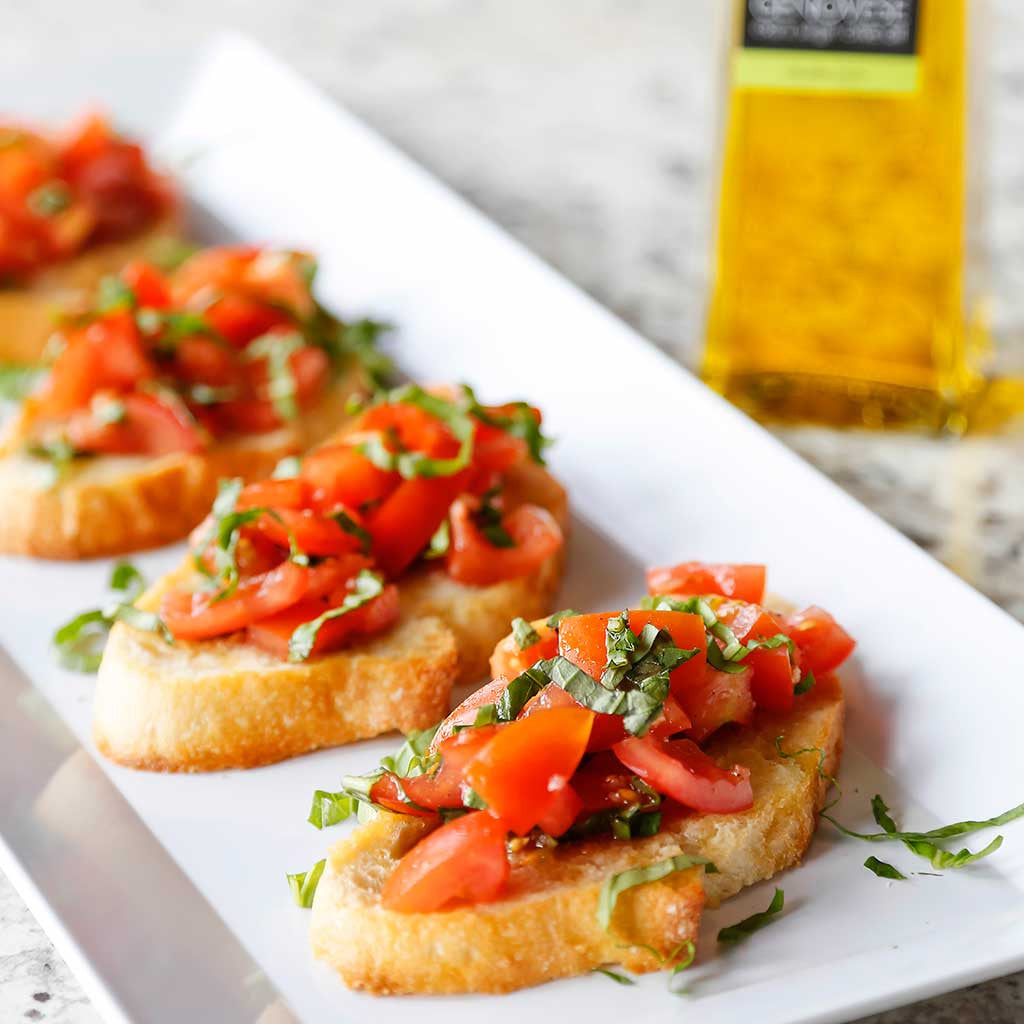 Tomato & Basil Bruscetta served on a white rectangular platter next to a bottle of Basil Genovese Infused Olive Oil on a marble counter top