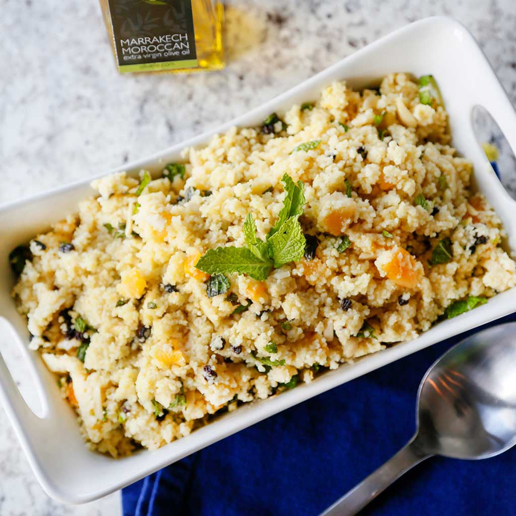 Moroccan Couscous Salad served in a white platter next to a bottle of Marrakech Moroccan Extra Virgin Olive Oil on a marble counter top.