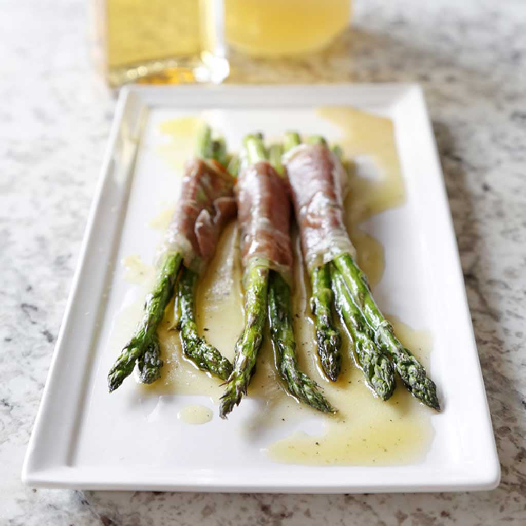 Pancetta-Wrapped Asparagus with Citrus Vinaigrette served on a white rectangular platter next to a bottle of White Balsamic Vinegar and Lemon Infused Olive Oil