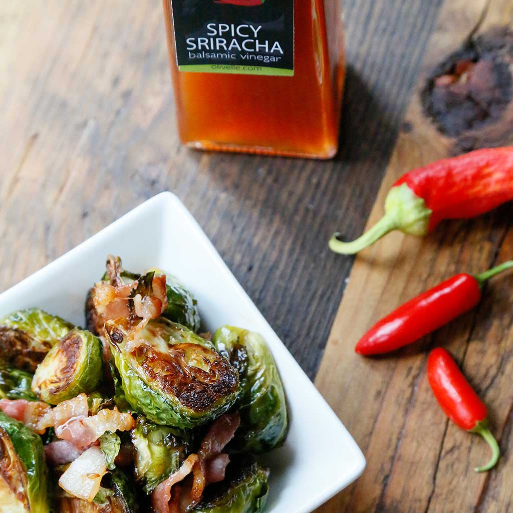 Bacon & Sriracha Roasted Brussel Sprouts served in a white square bowl next to a bottle of Spicy Sriracha Barrel Aged White Balsamic Vinegar and red chilis